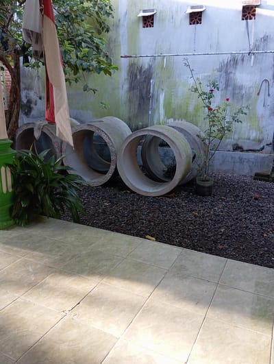 The image depicts an outdoor scene in what appears to be a backyard or side yard. In the foreground, there are several concrete pipes stacked on top of each other. Nearby are some plants. The ground is covered with gravel. A tiled surface is also visible. A wall is present with small square openings. In the background, there is a wall, stained and marked, with a single flowering plant. The location appears to be in Baron, Indonesia.