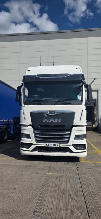 A front-facing medium shot captures a white MAN truck parked on a concrete surface marked with yellow lines. The truck, positioned centrally, faces the viewer directly. Its grille prominently displays the "MAN" logo, with the brand's lion emblem situated above it. The license plate reads "MJ75 HVY".
Behind the truck, a large, light-colored building with horizontal paneling forms the backdrop. To the left of the truck, a portion of a bright blue trailer is visible. The sky above is a vibrant blue, dotted with white, fluffy clouds. The lighting suggests it is daytime, with shadows cast on the ground. The scene appears to be in an industrial or commercial area.
The image was taken in Lichfield, United Kingdom.