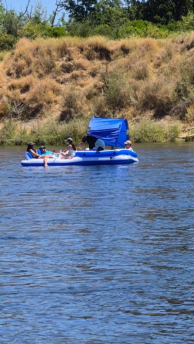 In the city of Rancho Cordova, United States of America, a blue and white inflatable raft with a blue canopy is floating on a body of water. Five individuals are present on the raft. Two individuals are seated on the left side, one wearing a dark bikini top and sunglasses, and the other in a blue shirt and cap. In the center, a person in a white top and cap is seated, looking at a phone. Towards the right, a person wearing a black shirt is partially visible, bent over the raft. Another individual in a white top and cap is seated on the far right. The raft is positioned in front of a dry, grassy hillside with scattered green foliage. The water is calm and reflects the blue sky.