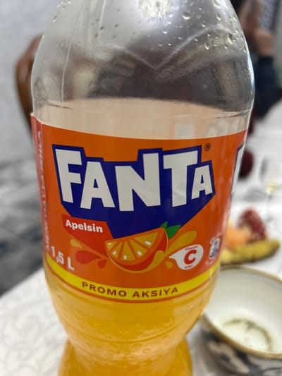 A close-up shows a bottle of Fanta orange soda. The bottle has condensation. The label is orange with "FANTA" written in large, blue letters. A slice of orange and the text "Vitamin C" are visible. "Apelsin" and "1,5 L" are also printed on the label. The word "Promo" is written vertically. The rest of the bottle is filled with yellow soda. A teacup is in the background.