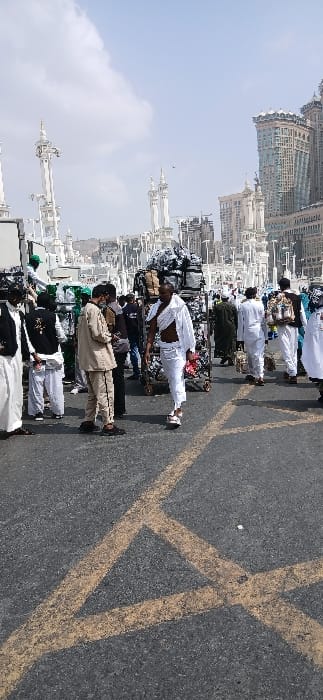 This image depicts a bustling outdoor street scene in Makkah Al Mukarramah, Saudi Arabia, under a partly cloudy sky. The foreground features dark asphalt pavement with faded yellow painted lines forming an intersecting pattern.
Approximately two dozen individuals are visible in various states of movement and interaction. In the mid-ground, slightly right of center, a dark-skinned male walks towards the viewer, clad in two white unsewn sheets of cloth (ihram garments), with a small white pouch secured around his waist. He appears to be barefoot or wearing light sandals.
To the left of the frame, several males are gathered near a white truck or large rectangular object. One male wears a tan uniform (trousers and jacket) and is looking downwards. Another male in a white thobe and a dark vest with white Arabic script is partially visible. Further to the right, behind the central figure in ihram, multiple large, multi-tiered metal carts, heavily loaded with bundled items (possibly merchandise or luggage covered in dark material), are being attended or moved by individuals.
In the background, a dense urban environment is present. Distinctive white minarets and classical Islamic architectural elements are visible, interspersed with modern, tall buildings, including a prominent high-rise structure with a teal facade on the far right, characteristic of the Abraj Al-Bait complex. A distant, arid hillside can be seen in the upper left background. A small bird is flying in the sky near the center-top of the frame.