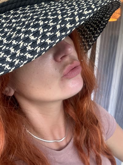 A close-up, low-angle medium shot shows the lower face and neck of a person wearing a wide-brimmed hat with a black and white woven pattern. The hat obscures the top of the person's face, but their lips are pursed in a kissing gesture. The person has fair skin and vibrant red hair, which is visible around their face and neck. They are wearing a pinkish-purple t-shirt and a thin, silver necklace. The background consists of vertical striped curtains or blinds. There are no discernable architectural elements or environmental features that would indicate a specific location within Saint-Claude, France.