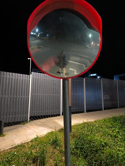 A convex mirror mounted on a pole reflects a person wearing a dark coat and a lighter colored top, standing on a sidewalk. The mirror is framed by a red border. Behind the mirror is a gray vertical slat fence. To the left of the fence is a light pole. To the right of the mirror, a neon sign with Cyrillic lettering is visible, indicating a business. The ground in front of the fence is a paved pathway, and to the right and foreground is a patch of green grass with small white flowers. The overall scene suggests an outdoor urban environment at night, illuminated by artificial lighting. The location is identified as Krasnaya Polyana, Russia.