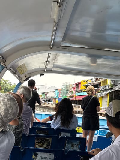 People are sitting on a boat, seemingly on a tour. The boat's white structure encloses them. Outside, there are colorful buildings along the water, possibly a tourist area. Some passengers are looking at the buildings, while others seem to be conversing. It seems to be a sunny day, and the boat is moving along the water.