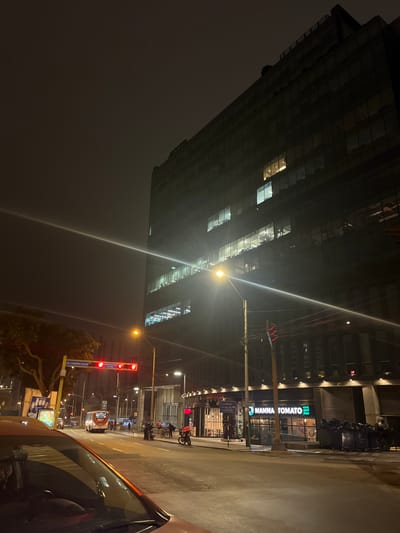 At night in Miraflores, Peru, a tall glass building dominates the scene, its upper floors disappearing into the fog. Streetlights cast a stark glow. The street is visible with a restaurant named "Mamma Tomato" with signage, trash bags stacked outside of the restaurant, motorcycles are parked along the sidewalk. The street is red with traffic lights and streetlights. A sign reads "AV. CASIMIRO ULLOA", indicating the street name. A car's side mirror and front window take up the right side of the frame, suggesting a view from inside a vehicle.