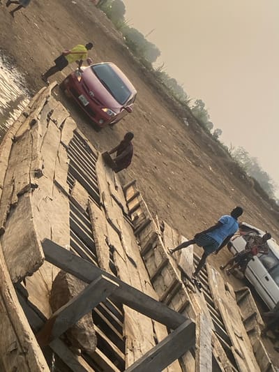 A tilted scene captures a red car positioned on a dirt incline near water. Several men are seen around what appears to be a large, dismantled wooden structure, possibly a boat under construction or repair. One man is leaning on the car, while others are walking or standing on the wooden planks. A white vehicle is visible further up the incline. The sky is overcast.