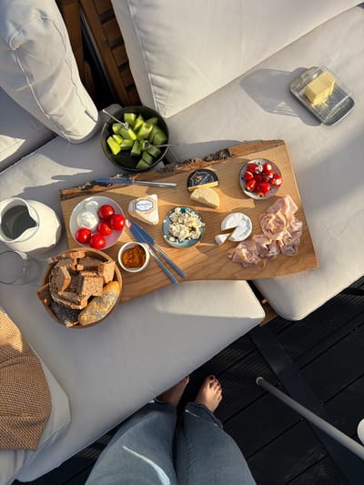 A high-angle, static shot captures a wooden board laden with food and drinks on an outdoor seating area. The board rests on a light-colored cushioned sofa, which is positioned on a dark wooden deck. In the foreground, a person's legs and feet, clad in blue jeans and wearing red nail polish, are visible, suggesting they are seated on the sofa.

The wooden board features an assortment of items: a wedge of cheese labeled "Peyrigoux," a round of brie cheese with a slice removed, a small plate of prosciutto, a small dish of what appears to be a spicy paste or dip, a bowl filled with mixed bread pieces, a plate with four cherry tomatoes and two white cheese balls, and a small blue dish containing crumbled white cheese mixed with red pieces. Adjacent to the board, a dark bowl holds cut green melon, with two forks inserted. A white pitcher and a glass carafe are also present on the sofa, to the left of the board. To the upper right of the scene, a small silver dish holds a block of butter. The lighting suggests it is late afternoon or early morning, casting long shadows across the sofa. The overall scene depicts a leisurely outdoor meal setting.