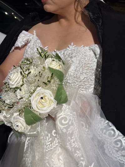 A bride is holding a bouquet of white roses adorned with small gemstones. She wears a white lace dress with a delicate floral pattern and a sheer veil. A black jacket covers her shoulders. The image suggests a formal event, possibly a wedding, given the attire and floral arrangement.