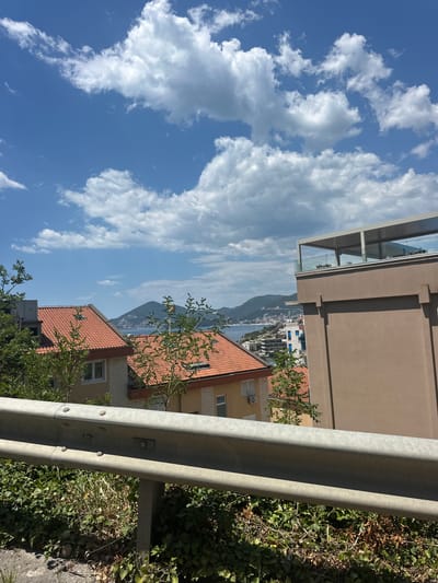 The image depicts a scenic view of the coastline in Pržno, Montenegro, with a clear blue sky filled with fluffy white clouds dominating the upper portion of the frame. In the foreground, a weathered metal guardrail runs horizontally across the image, separating a steep, green embankment covered in dense foliage from the rest of the scene. Behind the guardrail and embankment, two residential buildings with distinct orange-tiled roofs are visible, nestled amongst lush green trees. Further in the background, the azure waters of the Adriatic Sea stretch out towards a mountainous coastline dotted with more buildings and vegetation. A modern building with a rooftop terrace, featuring glass railings, stands prominently on the right side of the frame, partially obscuring the view of the distant landscape. The overall impression is one of a tranquil coastal town on a bright, sunny day.