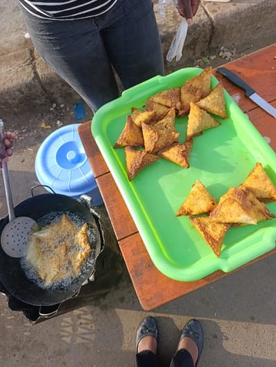 The image depicts an outdoor food preparation and selling scene in Kiuu ward, Kenya, captured from an overhead perspective. In the foreground, a person's feet, wearing dark flat shoes and dark trousers, stand on a concrete ground surface. To the left, a black frying pan sits on what appears to be a dark fabric or plastic sheet on the ground, containing several triangular pastries actively deep-frying in bubbling oil. A silver slotted spoon is partially visible, held by a hand, positioned inside the pan. A light blue plastic container with a lid is on the ground next to the frying setup.
To the right, a bright green plastic tray is placed on a brown wooden surface. This tray holds a significant quantity of cooked, golden-brown triangular pastries, arranged in multiple piles. A silver knife with a black handle rests on the wooden surface next to the tray. A person, visible from the waist down, stands behind the wooden surface, wearing dark jeans and a striped shirt. Their right hand holds a clear plastic bag. The concrete ground shows shadows consistent with outdoor lighting.