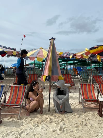 The image depicts a beach scene with numerous colorful beach chairs and umbrellas lined up on the sand. The sky is overcast with clouds, suggesting a cloudy day. In the foreground, a woman in a bikini is sitting on the sand, holding a smartphone and looking at it. Beside her, a person in a grey robe and head covering is also sitting on the sand, looking at their phone. Further back, more beach chairs and umbrellas are visible, along with a few other people. A small red flag is planted on a stick in the distance. The scene is set on a sandy beach, likely in a tropical location given the clothing and beach setup. The overall mood appears relaxed and casual, with people engaged with their phones.