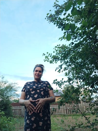 A pregnant woman stands outdoors in what appears to be a rural or suburban backyard. She is positioned centrally in the frame, facing the camera and smiling. Her hands are placed on her abdomen, forming a heart shape. She is wearing a dark, short-sleeved dress with a pattern of light-colored teddy bears. The background shows a wooden fence, some greenery, and a tree with green leaves obscuring part of the sky. The sky is light blue with some clouds. The presence of a wooden fence and what looks like a garden suggests a residential area, possibly in a village or on the outskirts of a town like Rusalevka, Russia, given the provided context. The lighting indicates it is daytime, possibly late afternoon or early evening.
