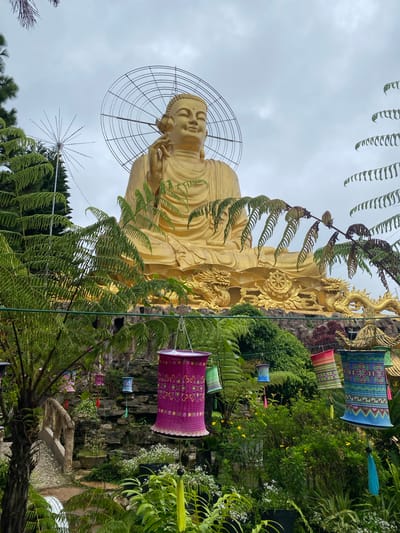 The image captures a large, golden Buddha statue seated outdoors in what appears to be a temple or garden setting in Lâm Viên - Đà Lạt, Vietnam. The Buddha is adorned with a circular halo or nimbus behind its head, and is depicted with its right hand raised, holding a flower. The statue is positioned on a rocky, tiered base, with ornate golden dragon sculptures flanking it. In the foreground, lush green ferns and other foliage create a natural frame for the scene. Several colorful, cylindrical lanterns, decorated with intricate patterns and tassels, hang from unseen supports, adding a festive touch to the environment. The sky is overcast, suggesting a cloudy day. The background reveals more greenery and architectural elements suggestive of a Buddhist sanctuary.