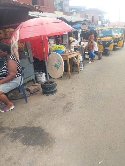 The image depicts a street scene in Agani, Nigeria, characterized by market activity along the left side of a paved road. In the foreground, a woman wearing a black and white striped top and light shorts is seated on a blue plastic chair, facing away from the camera. Her feet are adorned with white flip-flops. Adjacent to her are concrete blocks and two stacked black tires.
A red umbrella provides shade over a makeshift market stall, with a red banner partly visible beneath it, displaying "For You." A wooden table positioned to the right of the umbrella holds several plastic bowls containing yellow contents, alongside bottles and other small items. A large, circular, light-colored object with a green label leans against the table's leg. Another individual in a yellow top is partially visible behind this stall. The background on the left reveals various multi-story buildings with corrugated metal roofs, indicative of an urban setting.
The paved asphalt road, with a prominent dark stain in the lower-left foreground, extends toward the background. On the far right, three yellow motorized tricycles, commonly known as Keke Napep, are parked in a line. Several individuals are present near these tricycles; one person in a colorful patterned dress stands, while others are seated on small wooden stools. The overall lighting suggests daytime conditions.