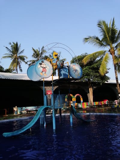 The image depicts a water park located in Kebonsari, Indonesia. In the foreground, a turquoise water slide curves into a dark blue swimming pool. The slide is supported by blue metal poles. Behind the slide, a multi-level structure, also painted blue, features circular panels adorned with cartoon characters: Mickey Mouse on the left and Donald Duck on the right, with the text "MUTIARA PARK" visible. Palm trees with green fronds are scattered throughout the background, with clear blue sky overhead. A dark building with a white corrugated roof is visible behind the water slide structure. Further back, a colorful fence made of vertical planks in various shades of red, yellow, green, blue, and orange encloses a seating area with tables and chairs, and small red and mushroom-shaped playhouses. Another archway structure with the word "WELCOME" is also visible. The scene appears to be set during the daytime, with bright sunlight illuminating the area.