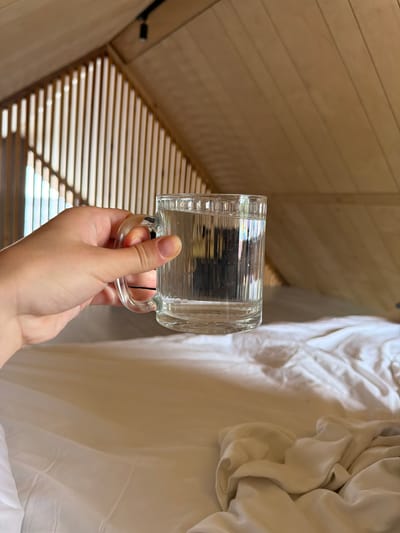 A hand holds a clear glass mug filled with clear liquid. The mug has vertical ridges and a curved handle. The background features a sloped wooden ceiling with visible planks and a black track light. To the left, a slatted wooden structure casts shadows on the ceiling. Below this, a white, rumpled bedsheet is visible. The scene is indoors and does not provide any information about the city of Семилуки, Russia.