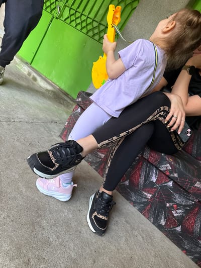 This image depicts three individuals—two adults and one child—in an outdoor or semi-outdoor environment in Kaspichan, Bulgaria, characterized by a concrete ground and a vibrant green metal gate or wall in the background.

On the right, an adult is seated on a dark fabric surface with a red and black abstract pattern. This adult wears a black top and black leggings featuring tan and black leopard print stripes along the sides. A gold-toned smartwatch is visible on their left wrist, and multiple rings adorn their fingers. Their right hand holds a smartphone encased in a black "Guess" branded cover. Their feet are clad in black sneakers with leopard print accents, one appearing to be a "Nike" Air Max style and the other a "Skechers" style.

Seated on the lap of this adult is a child, wearing a light purple t-shirt and light purple leggings, with pink "Champion" brand sneakers. The child holds a yellow toy guitar with a blue and yellow strap and looks upward and to the right. The child's left foot, in its pink sneaker, is resting directly on the top of the seated adult's left foot.

On the far left of the frame, the lower body of a third individual is partially visible, wearing black sweatpants and grey and white sneakers.