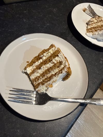 Two slices of layered cake, likely a sponge cake with cream filling, are presented on white plates. The cake on the left is a large triangular slice, featuring distinct brown cake layers alternating with thick white cream. A silver fork rests on the plate, with its tines piercing the edge of the cake slice. The plate itself is white with a thin dark rim. The second plate, partially visible in the upper right quadrant, holds a smaller portion of a similar cake, with a fork also present. Both plates sit on a dark, speckled countertop, possibly granite or a similar material. The lighting is dim, creating shadows and highlighting the textures of the cake and countertop. The scene appears to be an indoor setting, likely a kitchen or dining area, and there are no people visible. The presence of food and tableware suggests a meal or snack is being consumed. The specific location in Great Yarmouth, United Kingdom, cannot be determined from the visual information provided in the image.