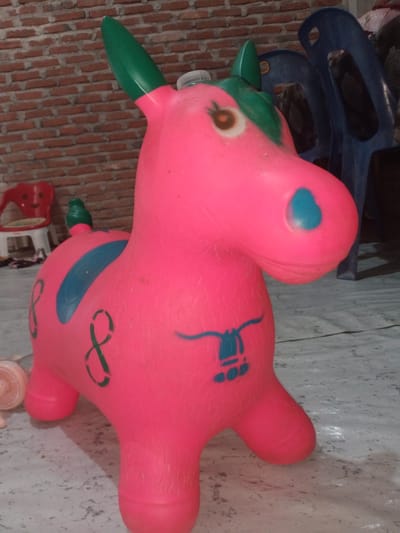 A pink inflatable toy horse with green ears and mane stands on a grey, speckled surface. The toy has blue markings on its body, including a stripe on its back and a horseshoe shape on its flank. In the background, a brick wall is visible, as well as a red chair and blue plastic chairs. The image appears to be taken indoors, possibly in the city of Cotseutui, Indonesia, as indicated by the presence of typical household items and building materials.