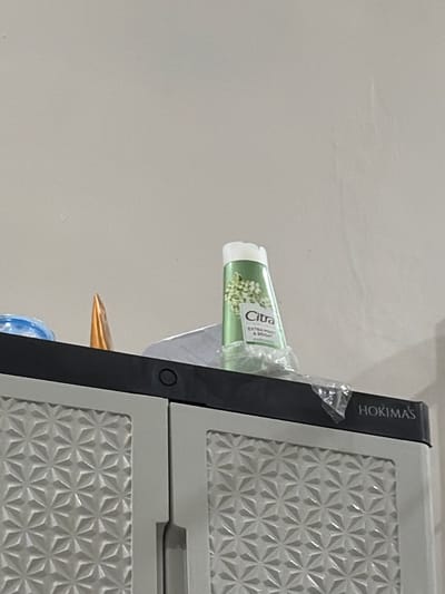 A Citra brand tube of lotion, possibly for skin care, sits on top of a light gray cabinet with a patterned design. The tube is green with white lettering and a white cap, and appears to be partially covered in clear plastic wrap. Next to the lotion tube, a small orange cone-shaped object is visible. The cabinet is labeled "HOKIMA'S" on the top right. The background is a plain, light-colored wall. The setting appears to be indoors, potentially a bathroom or storage area. This scene is not location-specific to Tupah, Indonesia, or any other city.