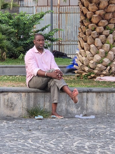 A Black man wearing a pink striped button-down shirt and khaki pants sits on a stone wall with his legs crossed. His left foot is bare, with the toes pointing upwards. He wears a silver bracelet on his left wrist and a ring on his right index finger. To his left, a pair of light blue flip-flops rests on the ground. To his right, a white plastic bag lies on the ground. In the background, a palm tree trunk stands to the right, and a metal fence and green bushes are visible behind the man. The scene appears to be in an outdoor public space, possibly a park or promenade.