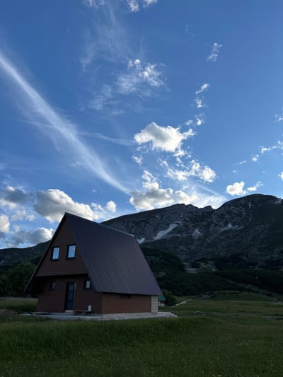 A triangular-roofed brown wooden cabin is situated in a grassy field with mountains in the background. The sky above is a vibrant blue with scattered white clouds. The cabin has two square windows on its upper level and a door on the front. A stone foundation is visible around the base of the cabin. The mountains are rocky and some patches of snow are visible. The surrounding landscape is covered in lush green grass. The scene appears to be in Virak, Montenegro.