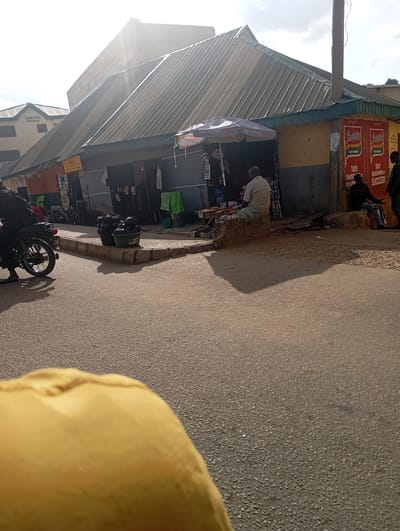In Kabong, Nigeria, a street scene unfolds under a bright sky with a few clouds. The image captures a row of shops with corrugated metal roofs, some with awnings. The shops appear to be selling various goods, with displays of items and signage visible.
In the foreground, a motorcycle is parked on the right side of the street, with its rider's leg visible on the left. Further down the street, a man sits on a raised concrete block in front of one of the shops, seemingly engaged in a transaction or waiting. Another man is seated on a similar block to his right, while a third person stands nearby.
Two large, dark green buckets labeled "HAPPY FAMILY" are placed on a concrete curb, suggesting they contain some sort of goods for sale. Adjacent to them are other items, possibly merchandise, arranged on the ground. The street itself is paved with asphalt, showing signs of wear and tear, with shadows cast from the buildings and objects. The overall impression is that of a busy marketplace or a commercial street in a Nigerian city.