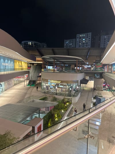 Here's a detailed description of the image:

**Setting:**

The image is taken at night within an upscale shopping mall, possibly in the city of Heroica Puebla de Zaragoza, Mexico. The location is a multi-level structure featuring modern architectural design with open spaces. The photograph is taken from an elevated position, possibly a balcony or upper level of the mall.

**Objects & Environment:**

*   Modern architectural elements are visible, including glass railings, concrete walkways, and high ceilings.
*   Various retail stores can be seen along the walkways, featuring brands like "Milamour" and "Pastini". The word "Puebla" can be seen on one of the posters.
*   There's advertising material visible, with one banner mentioning "MUY CERCA".
*   Escalators and stairs provide access to different levels of the mall.
*   Trees and landscaping elements can be seen in certain areas, adding greenery to the scene.
*   In the background, the lights of a residential or commercial building are visible.

**People & Actions:**

*   There are a few people walking along the walkways. The density of people is relatively low.

**Overall Impression:**

The image captures a nighttime scene in a modern, well-lit shopping mall. The architecture is sleek and contemporary. The atmosphere appears calm and relaxed.