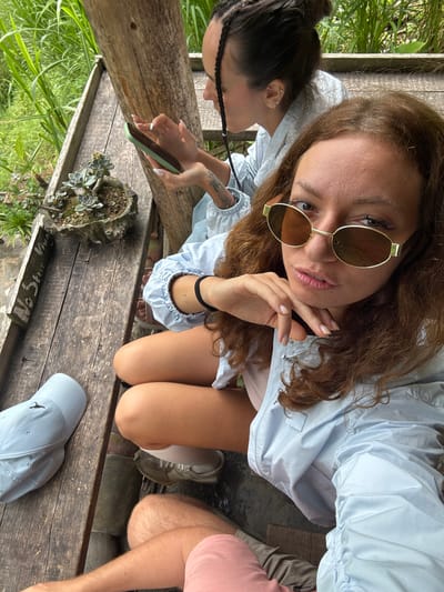 On a wooden platform in Yen Minh Commune, Vietnam, two women are the primary subjects of a high-angle shot. One woman, wearing sunglasses, is holding her hand to her face. The other is holding a phone. Both are wearing a pale blue jacket. One has hair in braids, while the other has longer, wavy brown hair. A potted succulent plant sits on the wood platform. A baseball cap and legs of another person are also visible on the platform. Greenery is visible in the background.