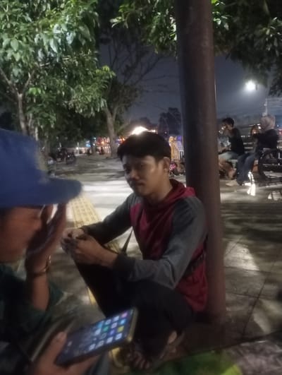 In Depok, Indonesia, at night, a group of people are gathered on a sidewalk near trees. The foreground features two individuals, one wearing a blue hat and the other a red and gray hooded jacket, both seated. The person in the hooded jacket is looking towards the left, with their hands resting on their lap. The other person, partially visible, is holding a smartphone. In the background, several other individuals are seated on benches, some of whom are looking at their phones. The scene is illuminated by artificial lights, creating a slightly hazy atmosphere.