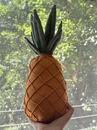 A hand holds a fabric pineapple against a background of out-of-focus green foliage visible through a screened window. The pineapple is orange with a crisscross pattern of darker thread and a green leafy top. The background suggests an outdoor environment with trees and dappled sunlight. The image does not contain any information that can be used to determine the location in the city of Chornomorske, Ukraine.