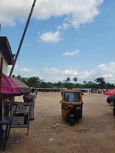 The image depicts an outdoor scene in Egbagu, Nigeria, characterized by a dusty, open area with scattered debris on the ground. In the foreground, a yellow and black auto-rickshaw (a TVS model) is parked. To the left, a sign for "ORBISON POS MONIEPOINT" is displayed, listing services like withdrawals, cash transfers, deposits, recharge cards, data, and bill payments, along with a phone number. Behind the sign, an umbrella with pink and brown fabric is partially visible. A person in a grey shirt and white trousers stands near the sign. Further in the background, a line of small shops or stalls with corrugated metal roofs are visible, some displaying signage such as "FRIENDS GLOBAL LTD" and "BAHWEBA COMPANY LTD". Several people are seen walking between the stalls. A black car is parked on the right side of the image. The sky above is a clear blue with scattered white clouds, and palm trees and dense green foliage form a backdrop behind the stalls. A large metal pole, possibly part of a utility structure, extends diagonally across the upper left portion of the image. The overall impression is of a busy, open-air market or commercial area in a rural or semi-urban setting.