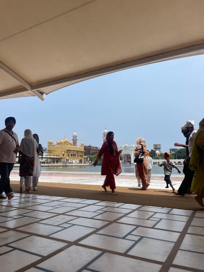 The image depicts the Golden Temple in Amritsar, India, viewed from under a large, light-colored awning. The foreground features a patterned marble floor. In the midground, several people are walking. On the left, a man wearing a pink shirt and a black turban is partially visible. Next to him, two women are dressed in traditional South Asian attire, one in white and the other in a maroon and white patterned outfit. Further to the right, a woman in a vibrant red outfit walks towards the left. A group of people are seen near the water's edge, including a child wearing a gray shirt and blue shorts, and several adults in traditional clothing and headwear. The iconic Golden Temple, a Sikh gurdwara, is prominently visible in the background, situated in the middle of a large body of water. The sky is clear and blue. The scene captures a moment of pilgrimage or tourism at this significant religious site.