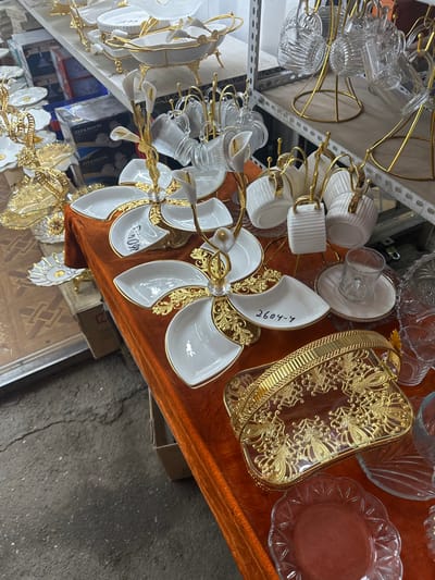 The image shows a stall in the Kara-Suu Bazaar, Kyrgyzstan, displaying a variety of decorative tableware. The items are primarily white ceramic with gold accents, including serving dishes, trays, and sets of cups and saucers. Some items feature floral designs or intricate gold filigree. The stall is set up on an orange velvet tablecloth, with shelves in the background containing more merchandise, including glassware. The ground in the foreground is a cracked, grey asphalt surface. There are no people visible in the frame.