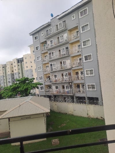The image displays an urban residential landscape in Ikate, Nigeria, under an overcast sky. The central focus is a multi-story apartment building, approximately seven to eight visible floors tall, with a light gray exterior, white window frames, and white horizontal metal balcony railings. Interspersed balcony walls are painted light yellow. Multiple windows are visible, some open, some closed. Several balconies feature clotheslines with garments hanging. Air conditioning units are attached to the building's facade. A blue satellite dish is mounted on the highest visible roofline. To the left and in the background, additional similar multi-story residential buildings, primarily in gray and beige tones, extend into the distance. In the mid-ground, a single-story structure with a light beige wall and a light gray pitched metal roof is visible. This structure is bordered by a green grassy area and a light beige concrete retaining wall, which separates it from the base of the taller apartment building. A dense green tree stands to the left, partially obscuring some background structures. A dark metal railing occupies the immediate foreground, suggesting an elevated viewing position. No individuals are clearly discernible in active roles.
