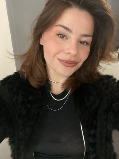 A light-skinned woman with brown hair and brown eyes smiles at the camera. She wears a black, sheer top and a fluffy black jacket. Two silver choker necklaces accent her neck. Her makeup is subtle, with a nude lip color and defined brows. The background appears to be a room with a light-colored wall.