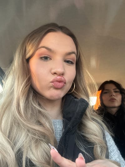 Two young women are shown in a selfie-style photograph. The main subject, in the foreground, has long, blonde, wavy hair, light skin, and is pouting her lips. She wears a dark-colored jacket over a light grey sweater and a gold hoop earring is visible. Her nails are long and decorated. Behind her, a second woman is partially visible, with dark hair and light skin, lying down. The background suggests an interior space, possibly a car, with a beige-colored ceiling. The location is not evident in the photograph.