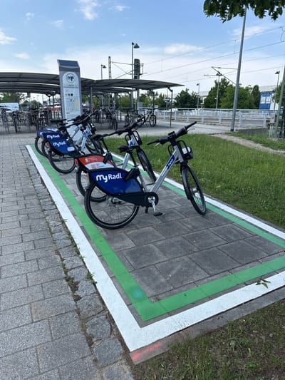 The image shows a row of rental bicycles parked at a station in Höhenkirchen-Siegertsbrunn, Germany. The bicycles are silver and blue and have "myRadl" branding on them. They are parked in a designated area marked with green and white lines on a paved surface. Behind the bicycles, there is a grassy area and some trees. In the background, there are overhead power lines, a railway station building, and a solar panel on a sign. The sky is blue with some clouds, suggesting it is daytime and the weather is fair. The scene appears to be a functional transportation hub, with the bicycles available for rent by commuters or travelers. The overall impression is one of modern urban infrastructure designed to facilitate sustainable transportation. No people are visible in the image.