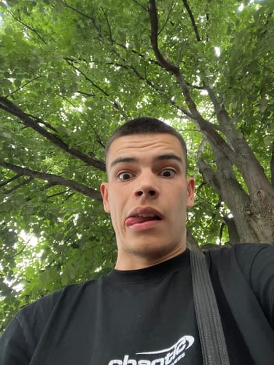 In a selfie taken from a low angle, a young man with short hair and wide, surprised eyes sticks his tongue out. He is wearing a black t-shirt with a logo that reads "Chaotic Fitness" visible on the lower portion of the shirt. The man is outdoors, with the lush green canopy of a tree filling the background. Sunlight filters through the leaves, creating a dappled effect. The image does not provide any specific clues about the location within Sopot, Bulgaria, other than it appears to be a natural, outdoor setting with significant tree cover.