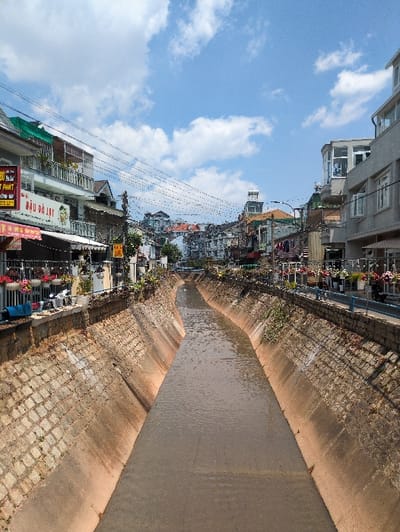 The image presents a concrete-lined canal, filled with murky brown water, cutting through an urban environment in Thành phố Đà Lạt, Vietnam. The canal extends straight into the distance, flanked on both sides by multi-story buildings, under a bright blue sky with scattered white clouds.

On the left bank, a row of buildings, primarily residential or mixed-use with ground-floor shops, features varied architecture, balconies, and numerous hanging flower planters. Overhead, a dense network of electrical wires is visible. A prominent white sign with green text reads "ĐẬU ĐÀ LẠT," and a partial sign indicates "PHAT." Awnings are extended over some shopfronts.

The right bank exhibits similar multi-story buildings, some with modern glass enclosures and balconies displaying drying laundry. A blue metal railing runs along the canal's edge, also adorned with hanging flower planters. Electrical wires are also present overhead on this side.

The canal walls are constructed with a lower concrete section and an upper layer of rough-hewn stone blocks, with sparse vegetation. In the background, more buildings ascend a hillside, and a notable structure with a peaked roof and a solar water heater is visible centrally, further down the canal's path where it passes under a distant bridge. No individuals, direct actions, or interactions are observable within the scene, which is illuminated by direct sunlight.