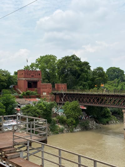 The image depicts a scene in Shuklaganj, India, focusing on the Ganges River and its surroundings. In the foreground, a metal walkway with stairs and railings extends over the murky brown water. Further back, a reddish-brown brick structure, possibly an old fort or tower with crenellated battlements, stands amidst lush green trees. A flag flies atop the structure. A rusty metal bridge, likely a railway bridge given its construction, spans the river, connecting the area to a landscape with more trees and some buildings on the far bank. The riverbank on the right is lined with accumulated debris, including plastic bags and other refuse, suggesting an urban or semi-urban environment. The sky is overcast with clouds.