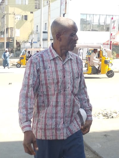 An adult male, appearing to be of African descent, is centrally positioned in the foreground of an urban street scene in Sabon Gari West, Nigeria, during daylight hours. He is shown in profile, facing towards the right side of the frame, with a closely shaved head and visible facial lines. He wears a long-sleeved shirt with vertical stripes in red, white, and dark blue/black, paired with dark blue trousers. His left hand is partially obscured by his clothing and holds a small white object.

In the mid-ground, a paved road features two yellow motorized tricycles. The closer tricycle on the right contains a driver wearing white attire and a cap. A second tricycle, further to the left, also contains a passenger. To the far left in the mid-ground, a person wearing a light blue robe and a light-colored cap is walking leftward.

The background consists of several multi-story buildings. On the right, a prominent white building with large windows displays a red "ZENITH" logo and "ATM" text, identifying it as a bank branch. To the left, a building with a yellowish facade is visible. Utility poles are present, and the ground in the lower right foreground shows scattered debris.