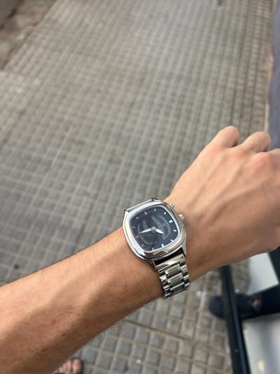 The image features a close-up view of a person's wrist wearing a silver watch with a black face. The watch has a distinctive cushion-shaped case and a metal link bracelet. The dial of the watch has white hour markers and hands, with a subtle pattern visible on the black surface.
The wrist is bare and has some light arm hair. The background consists of a textured gray surface, possibly a tiled or paved outdoor area, with a grid-like pattern. In the upper right corner, a dark, out-of-focus object is visible, potentially part of an outdoor structure or furniture. A sliver of a person's foot wearing a dark sandal is also visible at the very bottom right edge of the image.
The lighting suggests it is daytime, possibly sunny, as indicated by the bright illumination and some reflections on the watch. The overall scene appears casual and relaxed, possibly taken while the person is outdoors. The location context suggests Mérida, Spain. No visible text is present in the image.