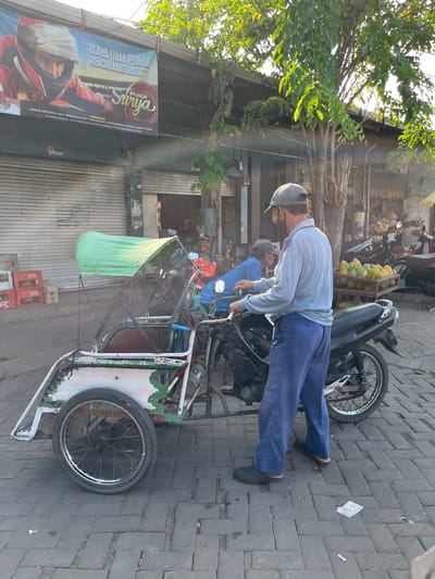 The image depicts a street scene in Juwet Kenongo, Indonesia. In the foreground, a man wearing a grey cap, a light blue long-sleeved shirt, and blue pants stands next to a three-wheeled vehicle that appears to be a modified motorcycle with a passenger carriage. The vehicle has a green canopy and white and green detailing. The man is facing away from the camera, looking towards the passenger carriage. He appears to be interacting with the vehicle or preparing to operate it. In the background, there are several other motorcycles, a fruit stand displaying papayas, and shops with closed roll-up doors. A banner is visible hanging above the shops with text in Indonesian, featuring an image of a person in a racing helmet. Trees with green foliage are also present, casting shadows on the scene, indicating it is likely daytime. The ground is paved with brick-like stones.