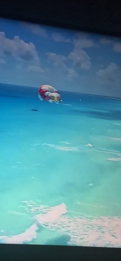 The image shows a parasail with a colorful canopy inflated and suspended in the air over a bright turquoise ocean. The parasail appears to be tethered to a boat, which is not fully visible but is suggested by the taut line extending from the parasail. There are faint white clouds in the blue sky above the water, and the ocean surface is calm with some lighter patches suggesting shallow water or underwater features. A dark, elongated object, possibly a boat or a wake, is visible in the water to the left of the parasail. The image is taken from an elevated perspective, looking down and across the water. There is no identifiable information within the image that would suggest a location in Bila Tserkva, Ukraine, as Bila Tserkva is an inland city and this image clearly depicts a coastal or oceanic scene.