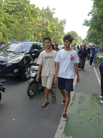 The image captures a vibrant outdoor scene on a street lined with trees, likely during the daytime. In the foreground, two young men walk towards the camera. The man on the left wears a light-colored short-sleeved t-shirt with the word "MARADUE" in a stylized font across the chest, light-colored shorts, and black slide sandals. He has dark hair and is looking slightly away from the camera. Beside him, to the right, is another young man wearing a white short-sleeved t-shirt featuring a red and black graphic logo (possibly a stylized "LA" or similar emblem), dark blue shorts, and black and white slide sandals. He has dark, wavy hair and is looking straight ahead.
Behind the man on the left, a black SUV (possibly a Hyundai Tucson) is visible on the road, with a black scooter in front of it. The scooter's license plate, "DR 4295 CJ 10.29," is clearly legible. The vehicles appear to be either stopped in traffic or moving slowly. Further in the background, a crowd of people, mostly young adults, can be seen walking on a sidewalk to the right of the road. Some individuals in the crowd are wearing face masks. On the far right, there are indications of market stalls or vendors, with green leafy items visible.
The street itself has a segment painted light green on the right side, suggesting a dedicated lane for pedestrians or cyclists. A decorative street light pole with an ornate golden top stands in the mid-ground, surrounded by lush green trees and foliage that canopy the street. The overall impression is of a busy public space, possibly a market day or a popular walking area, with a mix of pedestrian and slow-moving vehicle traffic. The lighting suggests it is daytime, with bright ambient light and no harsh shadows, indicating either an overcast sky or early/late afternoon.