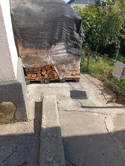 A high-angle, eye-level outdoor shot captures a pile of firewood covered by a black tarp and clear plastic sheeting. The firewood is neatly stacked on wooden pallets. To the left of the firewood, a portion of a building wall with a textured finish is visible. The ground is concrete with cracks and some light-colored stones embedded. To the right of the firewood, a metal fence runs horizontally, separating the paved area from a grassy area with trees. The sun casts shadows on the ground. The scene appears to be in a residential area.