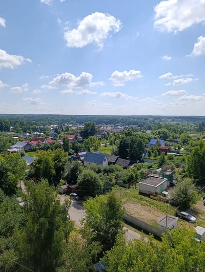 This is an aerial, eye-level, medium shot of a semi-rural Russian village on a sunny day with a few scattered clouds. The scene is dominated by a dense canopy of lush green trees interspersed with houses that feature red, gray, and blue roofs. Some of the houses have visible yards and fences. In the foreground, there are more trees and a dirt road with two cars parked on it. The sky is a vibrant blue with white, puffy clouds, suggesting it is daytime, likely late morning or early afternoon given the bright sunlight. The overall impression is one of peaceful, quiet, everyday life in a small Russian village, likely in Alexandrov, Russia, as per the location context. There is no visible text in the image.