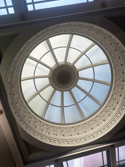 The image displays an upward view into the interior of a building, centered on a large, circular glass dome or skylight. The dome features a white-painted radial and concentric framework, segmenting the glass into numerous panes, with a smaller, dark circular element at its apex. This skylight is encircled by an elaborate, off-white decorative plasterwork band, intricately carved with repeating foliate motifs and accented with small, sparsely distributed gold-colored details. Beyond this ornate ring, the ceiling structure includes a darker, patterned molding or cornice running along an architectural recess. Additional rectangular skylight sections with gridded frames are visible at the top and bottom of the frame, hinting at a larger glass ceiling structure. The glass panes of the main dome transmit varying light, appearing from bright white to pale blue, suggesting natural daylight and a partially clear or lightly clouded sky. This architectural feature is located within the former Post Office building, now a retail establishment, on High Street in Salisbury, United Kingdom.