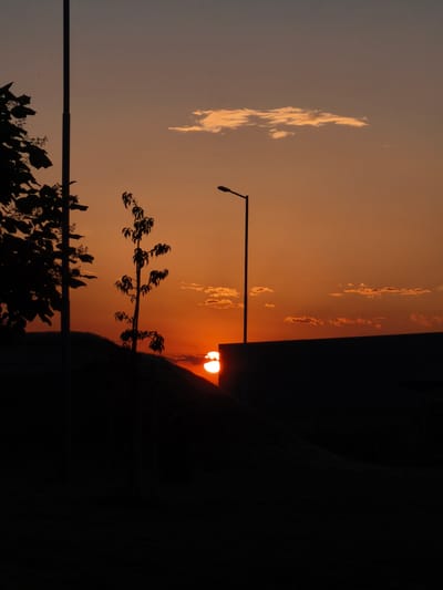 A solitary lamppost stands silhouetted against a vibrant orange sunset sky in Benkovski, Bulgaria. In the foreground, the dark shapes of trees and a gently sloping hill are visible. The setting sun, a bright orb, is partially obscured by a dark, rectangular structure. Small clouds drift across the sky, tinged with the warm hues of the sunset.
